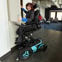 A Caucasian woman wearing a dark grey suit and a royal blue mock neck shirt uses her Corpus F5 power chair. It is in the ActiveReach position and she is reaching into a high cupboard while smiling at the camera. A living room area is behind her. thumbnail