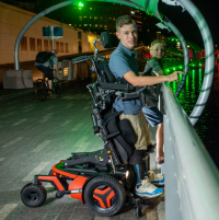 A young man using an F5 Corpus VS standing wheelchair holds his smaller brother up to the railings of a bridge to view the river below. It's nighttime and they are both smiling towards the camera. thumbnail