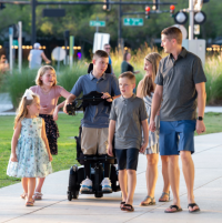 Landon, a young Caucasian man, uses his Permobil F5 VS standing wheelchair while rolling with his family in a park. They are on a cement pathway with tall grass and a street scene in the background. His family are looking at him and smiling. thumbnail