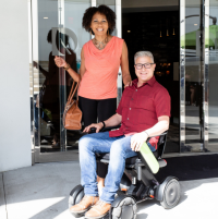 A man sits in a white WHILL C2 power chair with a woman standing beside him. They are both smiling at the camera. thumbnail
