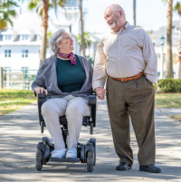 A senior couple strolls hand in hand down a path dappled with sunlight. The woman uses a WHILL power chair. They are smiling at each other. thumbnail