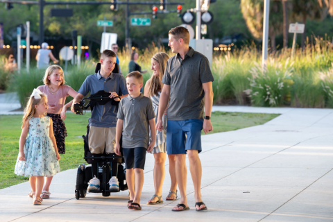 A young person in a standing power chair wheels along a path with their family.